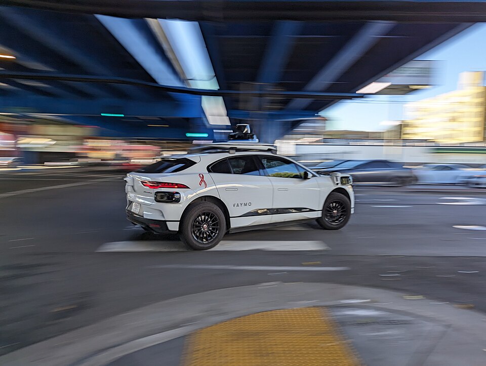 A white Waymo robotaxi with a roof-mounted sensor pod crosses an intersection under an overpass, sharp against blurred traffic and buildings.